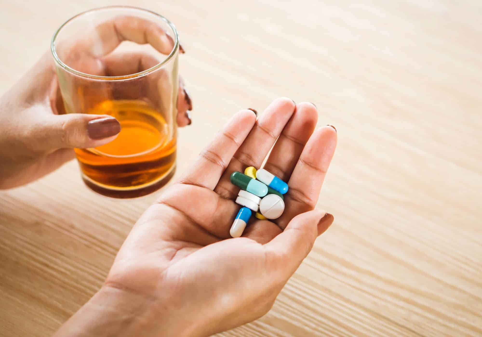 The image depicts a close-up, top-down view of a person’s hands positioned on a light-colored wooden surface. On the left side, a hand holds a short, clear glass filled with a golden-amber liquid, likely whiskey or another alcoholic beverage. The glass is simple in design, allowing the color of the drink to be the focal point. The hand holding the glass has a neatly manicured, dark-colored nail polish. To the right of the glass, the other hand is open, palm up, and contains a collection of various pills and capsules. There are approximately seven pills visible, in a range of colors including white, yellow, blue, and green. Some are capsules, appearing elongated with colored bands, while others are traditional tablet shapes. The skin tone of the hands suggests a person of color. The overall composition creates a stark contrast between the liquid in the glass and the solid medication in the palm, potentially symbolizing a difficult choice or a dangerous combination. The lighting is soft and natural, highlighting the textures of the skin, wood, and glass. The image evokes themes of health, addiction, self-medication, or the conflict between coping mechanisms. The shallow depth of field keeps the focus on the hands and their contents, blurring the background and emphasizing the central subject matter.